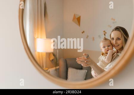 Glückliche Mutter, die eine kleine Tochter trägt und zu Hause im Spiegel reflektiert Stockfoto