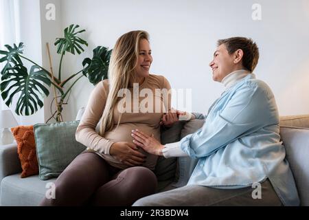 Seniorin mit schwangerer Tochter, die zu Hause auf dem Sofa sitzt Stockfoto