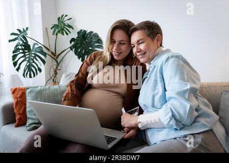 Lächelnde Frau, die mit ihrer schwangeren Tochter zu Hause online einkauft, über ein Notebook Stockfoto