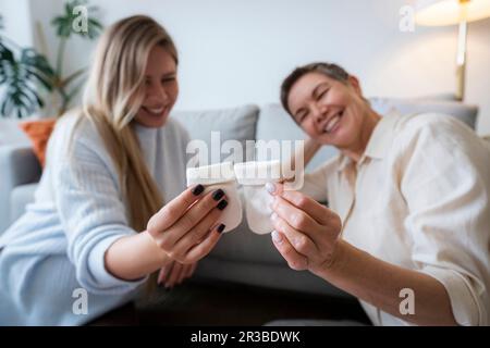 Mutter und schwangere Tochter halten Babysocken, die zu Hause sitzen Stockfoto