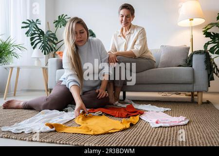 Mutter und schwangere Tochter sorgen zu Hause für Babykleidung auf dem Boden Stockfoto