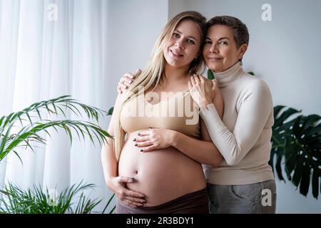 Lächelnde ältere Frau, die schwangere Tochter umarmt, die zu Hause steht Stockfoto