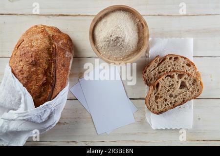 Holztisch mit einem Sauerei-Brot, zwei Scheiben Brot und etwas Mehl. Stockfoto