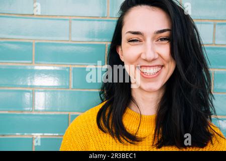 Lächelnde junge Frau mit schwarzem Haar vor einer Ziegelwand Stockfoto