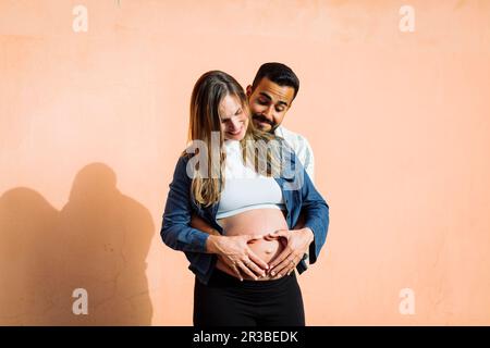 Lächelnder Mann, der den Bauch einer schwangeren Frau vor der Wand herzförmig macht Stockfoto