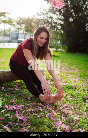 Lächelnde schwangere Frau, die Stretching-Übungen macht und auf dem Rasen sitzt Stockfoto