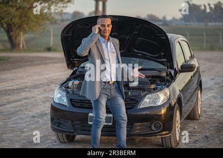 Ein junger Latino in einem Anzug, der vor seinem Auto telefoniert und am Straßenrand eine Panne hat. Stockfoto