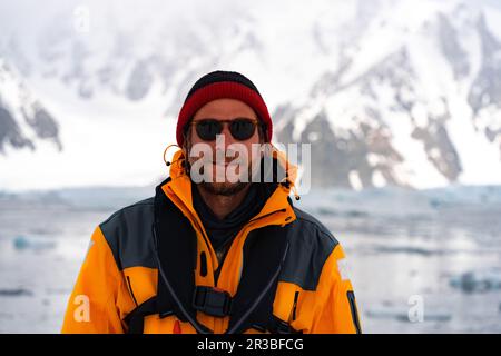 Junger männlicher Tourist mit gelber Jacke in der Antarktis, lächelnd. Eisberge im Hintergrund. Wandern mit Rucksack in den Bergen, in der Antarktis Stockfoto