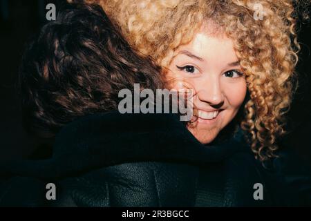 Lächelnde junge Frau mit lockigem Haar, die eine Freundin umarmt Stockfoto