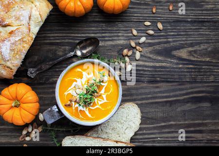 Ein Flatlay frischer Kürbissuppe garniert mit saurer Sahne, gerösteten Kürbiskernen und Thymian mit hausgemachtem Brot im Hintergrund. Stockfoto