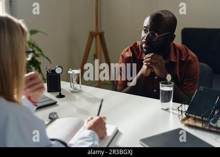 Aufmerksamer Patient mit gefalteten Händen sitzt mit einem Psychologen in der Praxis Stockfoto