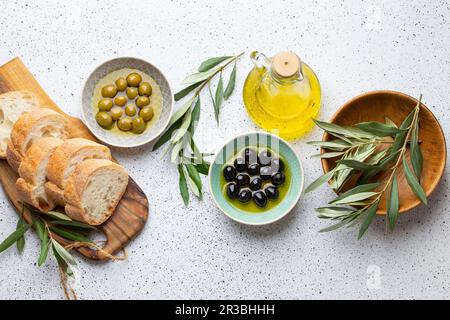 Grüne und schwarze Oliven mit Olivenöl in einer Glasflasche, Olivenbaumzweige und geschnittenes frisches Ciabatta-Brot Stockfoto