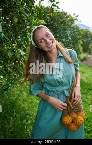 Lächelnde Frau mit Orangen in einem Netzbeutel im Obstgarten Stockfoto