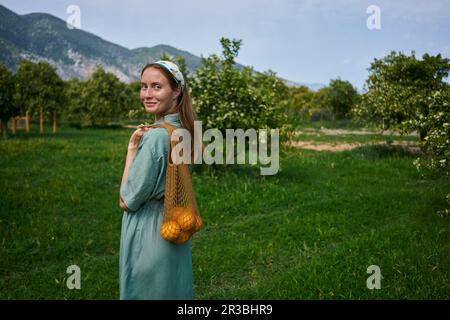 Lächelnde Frau mit Orangen in einem Netzbeutel, die auf Gras im Obstgarten steht Stockfoto