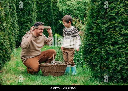 Der Sohn steht neben dem verspielten Vater und hält Baumstämme im Garten Stockfoto