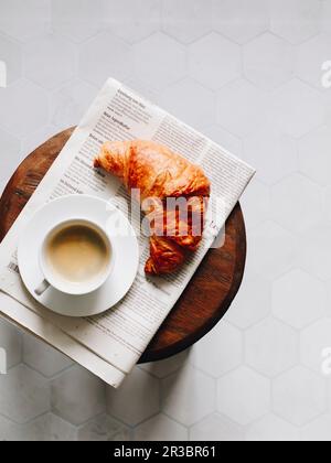 Frühstückscroissant, Tasse Kaffee und Zeitung auf einem Holzhocker Stockfoto