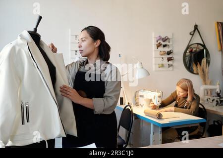 Junge brünette Frau, die an der Schaufensterpuppe steht, mit einer neuen weißen Lederjacke gegen einen reifen Kollegen, der Kleidung an eine Maschine näht Stockfoto