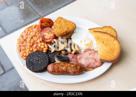 Traditionelles englisches Frühstück mit gebackenen Bohnen, gegrillten Tomaten, Kartoffelrösti, Spiegelei, Toast, Speck, Pilze, Wurst, Pudding Stockfoto