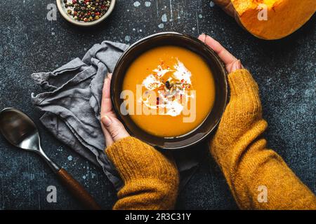 Weibliche Hände in gelbem Strickpullover mit einer Schüssel mit Kürbiscreme-Suppe Stockfoto