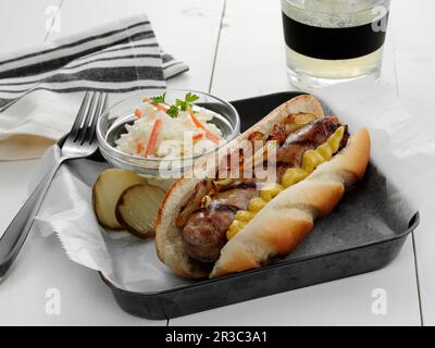 Eine gegrillte Bratwurstwurst auf einem Brötchen mit gelbem Senf und sautierten Zwiebeln, serviert mit Krautsalat und Gurken Stockfoto