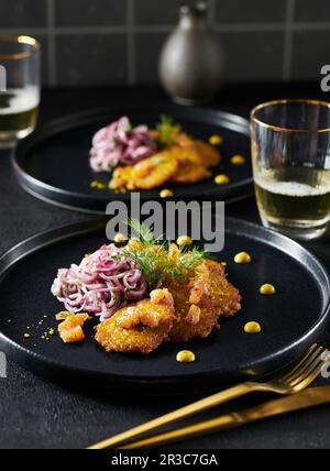 Kleine Lachskuchen mit Zwiebelsalat auf einem schwarzen Teller, mit goldenem Besteck Stockfoto