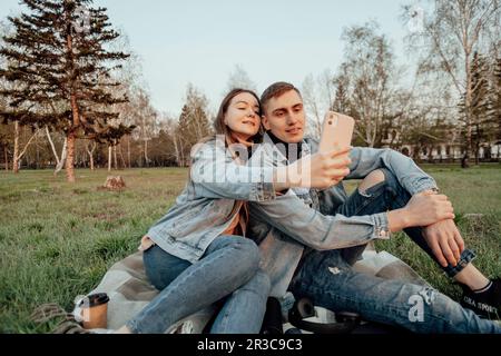 Ein junges, modisches Paar lächelt, während es im Park auf das Telefon schaut, Selfie Stockfoto
