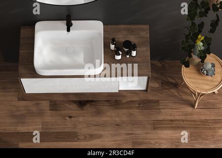 Blick von oben auf einen Schminktisch aus Holz mit weißem Keramikwaschbecken und einem modernen Wasserhahn in einem Badezimmer mit Morgensonnenlicht und Schatten. Leerzeichen für Stockfoto