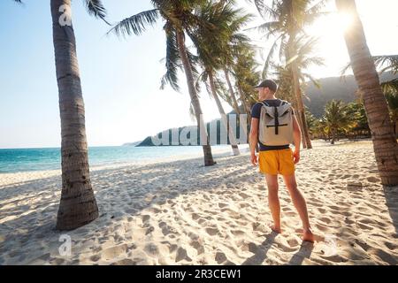Rückblick auf Touristen mit Rucksack, der auf einem wunderschönen Sandstrand mit Palmen spaziert und den Sonnenuntergang betrachtet. Cham-Inseln, Vietnam. Stockfoto