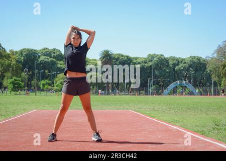 Junge lateinin argentinischer ethnischer Herkunft in schwarzer Sportbekleidung, steht draußen, streckt Arme und Schultern vor dem Training, schaut in die Kamera, CO Stockfoto