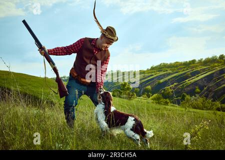 Porträt mit einem Mann, alter Jäger mit Retro-Hut und Federwaffe und streicheln den Hund englischer springer Spaniel über die wunderschöne Natur Stockfoto