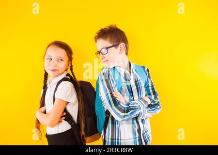 Der Junge und das Mädchen der Grundschule stehen auf gelbem Hintergrund. Die Pupillen stehen mit gekreuzten Händen und sehen sich an Stockfoto