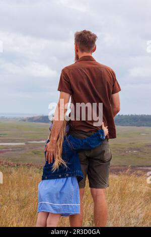 Vater und Tochter stehen auf einem Hügel mit Blick auf die Natur. Kleines blondes Mädchen mit Umarmung mit Dad draußen. Liebevolles Kind, umarme ihren Dadd Stockfoto