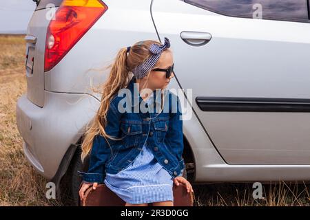 Mädchen entspannt und genießt den Roadtrip am silbernen Auto. Ein glückliches Kind reitet auf das Abenteuer zu. Süße kleine Dame in Jeansjacke, schwarze Sungla Stockfoto