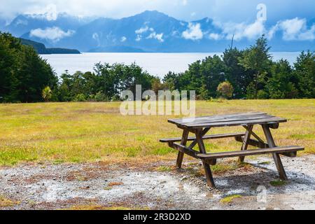 Picknicktisch aus Holz über dem malerischen Manapouri Lake und den Bergen des Fiordland National Park in Southland, South Island von Neuseeland Stockfoto