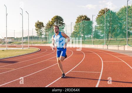 Junger Mann, ein Profisportler mit einem ernsthaften und fokussierten Gesicht, der auf einer Outdoor-Sportstrecke in der Nähe des Sportplatzes läuft Stockfoto