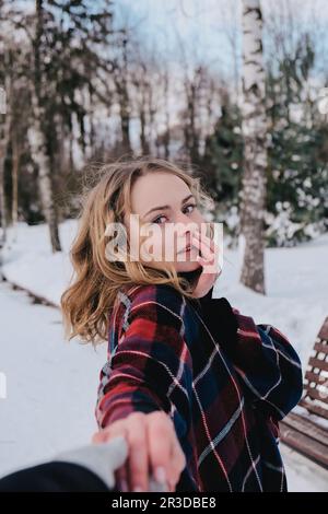 Eine lockige blonde, hübsche junge Frau in einem Poncho im Winterpark. Kaltes Wetter. Urlaubs-, Saison- und Freizeitkonzept. Weihnachten, Neujahr Stockfoto