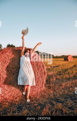 Porträt einer jungen, glücklichen Frau, die ihren Hut auf dem Heuhaufen im Morgensonnenlicht, auf dem Land fliegt. Eine schöne Frau in einem Kleid sitzt auf einem Stockfoto