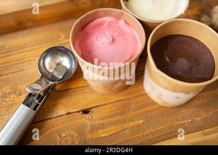 Eiscreme in einer Schachtel mit verschiedenen Geschmacksrichtungen steht auf einem Holztablett zusammen mit einer Eiskrem-Schaufel aus Metall Stockfoto