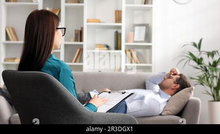 Ein junger Mann liegt auf einer Couch bei einem Psychologen-Termin. Eine brünette Brillenpsychologin führt eine psychologische Sitzung durch Stockfoto