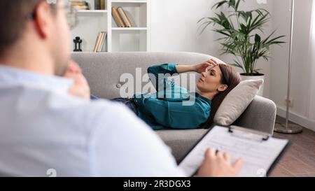 Eine junge Frau liegt auf einer Couch beim Termin eines Psychologen. Ein brauner, männlicher Psychologe mit Brille führt eine psychologische Sitzung durch Stockfoto