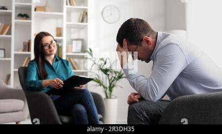 Ein professioneller Psychologe konsultiert einen männlichen Patienten in einem Zustand von Stress und Depression, der auf einem Stuhl im Büro sitzt. Sie schreibt auf Papier Stockfoto