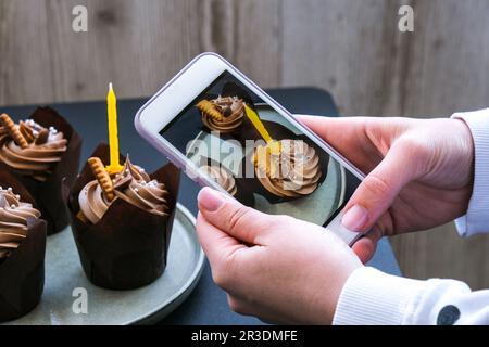 Weibliche Hände fotografieren köstliche Schokoladen-Cupcakes mit Sahne auf dunklem Hintergrund. Influencer drei Schokoladen-Muffins. Birt Stockfoto