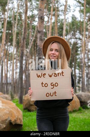 Porträt einer jungen Weißen mit Hut, die Pappe in der Hand hält, mit dem Text, DEN WIR RAUSGEHEN wollen. Naturhintergrund. Protester AC Stockfoto