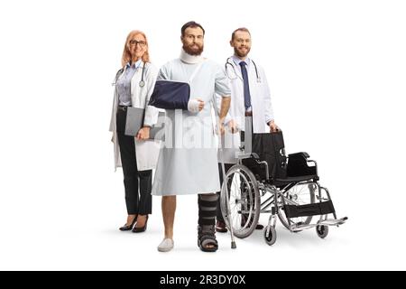 Bärtiger Mann in einem Krankenhauskleid mit gebrochenem Bein und Arm stehend mit einem Team von Ärzten isoliert auf weißem Hintergrund Stockfoto