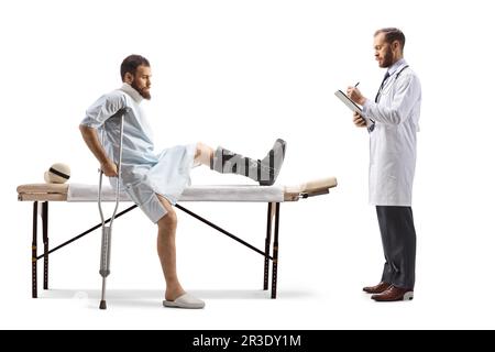 Verletzter Mann mit orthopädischem Stiefel und Halskragen, der auf einem Therapietisch sitzt und ein Therapeut, der ein isoliertes Dokument auf weißem Hintergrund schreibt Stockfoto