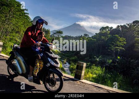 Sleman, Yogyakarta, Indonesien. 23. Mai 2023. Eine Frau, die mit ihrem Kind Motorrad fährt, überquert eine Brücke mit Mount Merapi im Hintergrund in Sleman, Yogyakarta, Indonesien, Dienstag, 23. Mai, 2023. Das Forschungs- und Entwicklungszentrum für Geologische Katastrophentechnologie (Geological Disaster Technology Research and Development Center, BPPTKG) hat 26 glühende Lavalawinen mit einer maximalen Gleitrate von 2000 Metern westlich des Südwestens oder des Flusses Bebeng erfasst. (Kreditbild: © Slamet Riyadi/ZUMA Press Wire) NUR REDAKTIONELLE VERWENDUNG! Nicht für den kommerziellen GEBRAUCH! Stockfoto