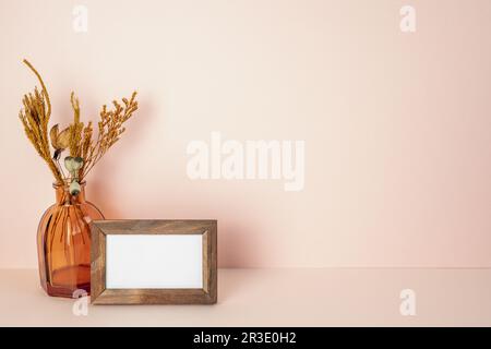 Holzbilderrahmen mit leerer Karte, braune Vase mit trockener Blume auf pinkfarbenem Tisch. Rosafarbener Wandhintergrund. Speicherplatz kopieren Stockfoto