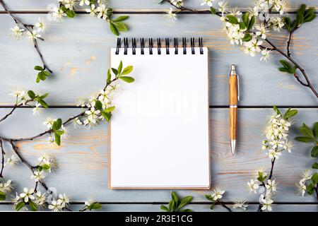 Öffnen Sie das Notizbuch mit leerer Seite und den Stift auf einem blauen Holztisch mit Kirschblüten. Draufsicht, flach liegend, Arbeitsbereich auf blauem Hintergrund. Hallo Spri Stockfoto