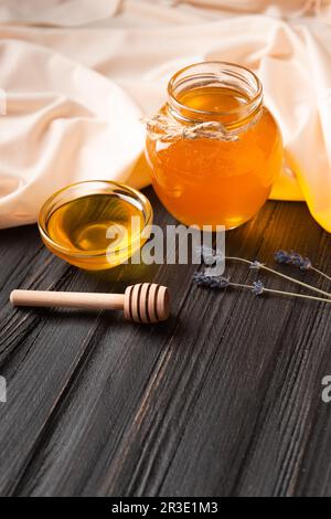 Honig in einem offenen Gefäß und einer transparenten Schüssel, einem Holzdipper, Lavendelblumen auf gelbem Textil und dunklem Holzhintergrund. Stockfoto