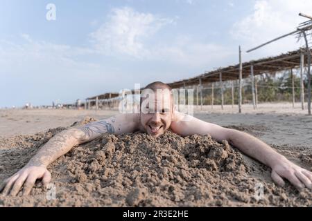 Glücklicher junger Kaukasier, der den Strand genießt, auf dem Sand liegt und vor der Kamera lächelt Stockfoto